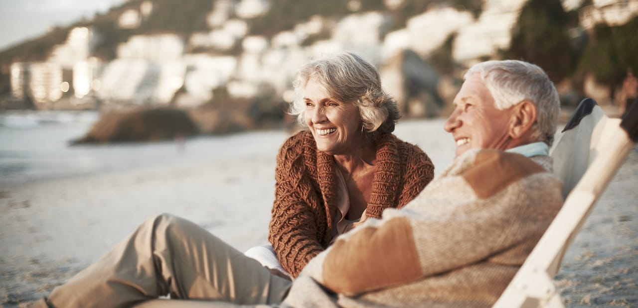 Retired couple laughs while lounging on a beach. 